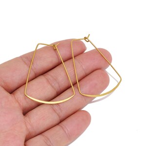 May include: A pair of gold-toned hoop earrings with a trapezoidal shape. The earrings feature a thin, wire-like metal construction with a curved bottom edge and a simple post-style closure mechanism at the top of each frame.