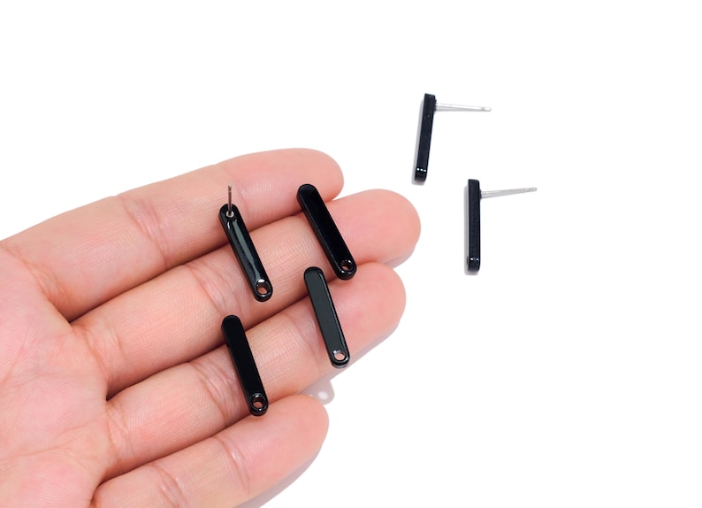 May include: A collection of small, glossy black bar-shaped stud earrings. Each earring features a smooth, elongated rectangular design with a rounded bottom edge and a small circular hole at the base, paired with a thin metal post for attachment.