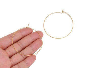 May include: A pair of thin, circular gold-toned hoop earrings. One earring is held in the palm of a hand, while the other is positioned separately to the right. Both hoops feature a simple, minimalist wire construction with a small post closure.