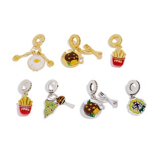 May include: A selection of food-themed charms. The charms depict a fried egg, a plate of food with cutlery, a container of chips, a slice of pizza, a dessert, and a plate of food with cutlery. The charms are gold and silver.