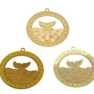 May include: Three gold-colored, circular pendants with a whale tail design. Each pendant features a whale tail emerging from stylized waves within a decorative circle. The pendants have a small hole at the top for hanging.