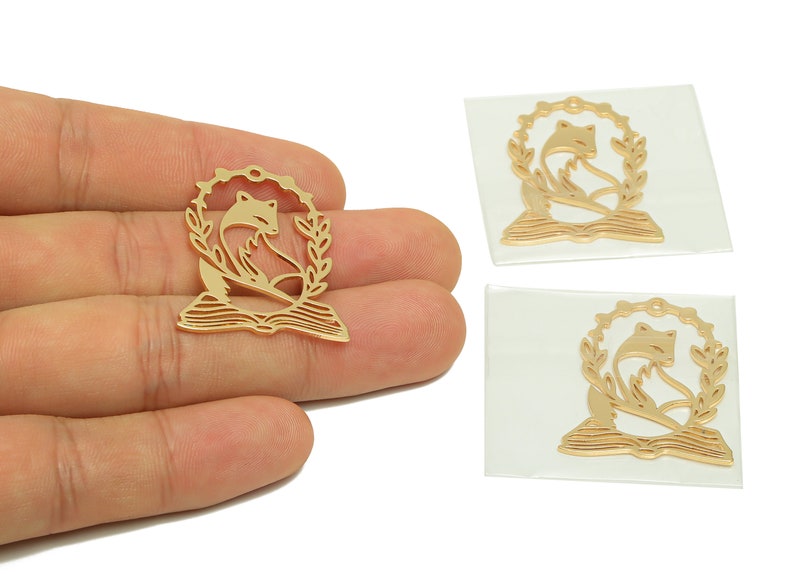 Pode incluir: Tr&ecirc;s pingentes de metal dourado com uma raposa sentada em um livro com uma coroa de louro ao redor. Os pingentes est&atilde;o sobre um fundo branco.