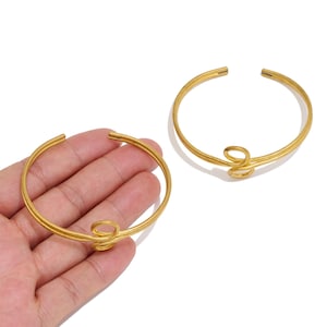 May include: Two gold-colored metal cuff bracelets, each featuring a smooth, circular band design with a central decorative loop detail. The bracelets have an open-ended construction, allowing for adjustment when worn on the wrist or arm.