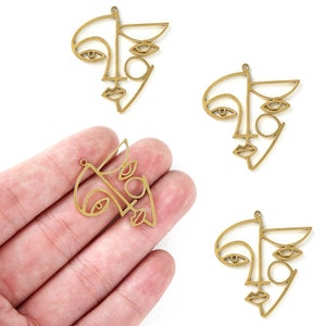 K&ouml;nnte beinhalten: Drei goldfarbene Metall-Anh&auml;nger in Form von abstrakten Gesichtern. Jeder Anh&auml;nger zeigt eine stilisierte Umrandung eines Gesichts mit zwei Augen, einer Nase und einem Mund.