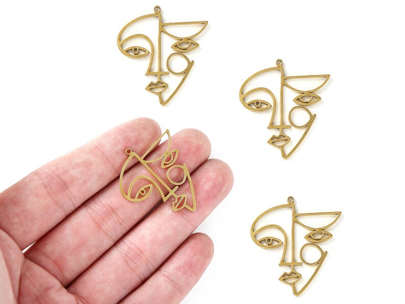 K&ouml;nnte beinhalten: Drei goldfarbene Metall-Anh&auml;nger in Form von abstrakten Gesichtern. Jeder Anh&auml;nger zeigt eine stilisierte Umrandung eines Gesichts mit zwei Augen, einer Nase und einem Mund.