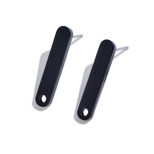May include: A pair of black stud earrings featuring a long, rectangular bar shape with rounded ends and a small circular cutout at the bottom. Each earring includes a silver-toned metal post attached to the back for secure placement.