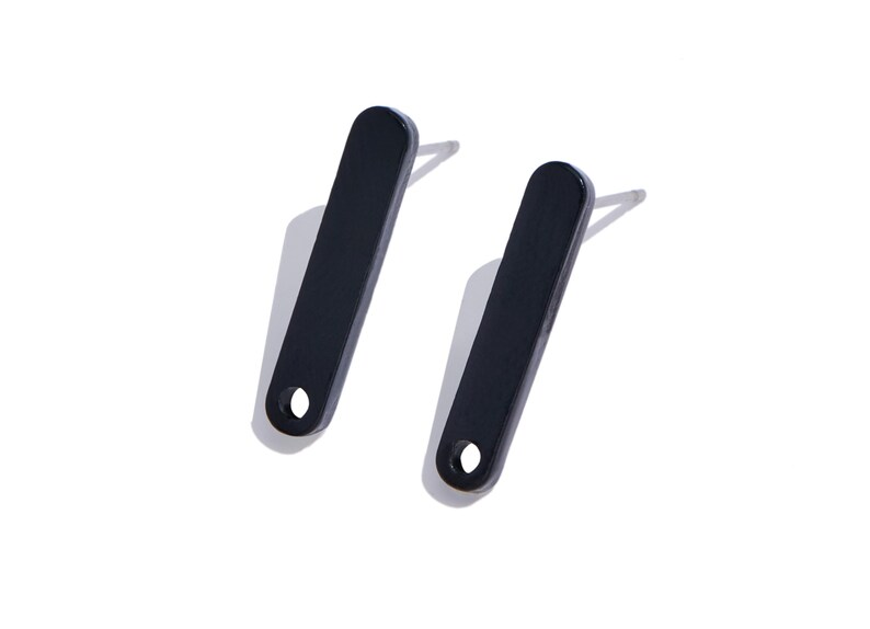 May include: A pair of black stud earrings featuring a long, rectangular bar shape with rounded ends and a small circular cutout at the bottom. Each earring includes a silver-toned metal post attached to the back for secure placement.
