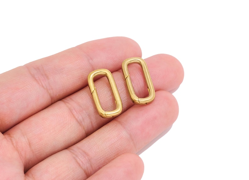 K&ouml;nnte beinhalten: Zwei ovale Federringe aus goldfarbenem Metall mit einer glatten, polierten Oberfl&auml;che. Jeder Ring verf&uuml;gt &uuml;ber einen Klappmechanismus an einer Seite, wodurch sich die Schlaufe &ouml;ffnen und sicher schlie&szlig;en l&auml;sst, um Gegenst&auml;nde zu verbinden.