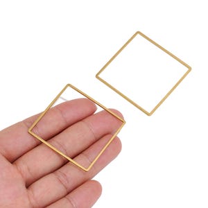 Könnte beinhalten: Zwei dünne, quadratische Metallrahmen mit mattem Goldfinish. Jeder Rahmen besteht aus vier geraden, schlanken Stäben, die an den Ecken zu einer hohlen, offen gestalteten geometrischen Form verbunden sind. Das Metall hat eine glatte Textur.