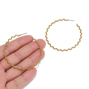 May include: A pair of large hoop earrings featuring a distinct wavy, rippled texture throughout the circular frame. The earrings are crafted from a matte, antique gold-toned metal and include a standard post-back closure mechanism for secure wear.