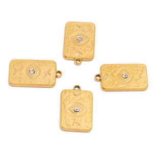 Pode incluir: Quatro pingentes retangulares dourados com um design de olho e um pequeno cristal transparente no centro. Os pingentes têm uma superfície texturizada com detalhes florais e de lua crescente. Cada pingente tem um pequeno laço para fixação.