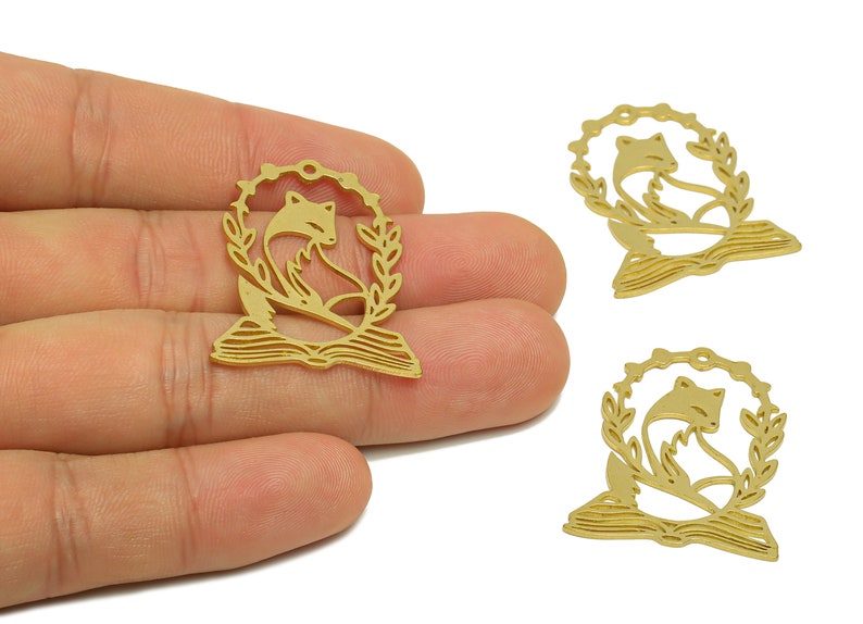 Pode incluir: Tr&ecirc;s pingentes de metal dourado com uma raposa sentada em um livro aberto com uma coroa de folhas ao redor.