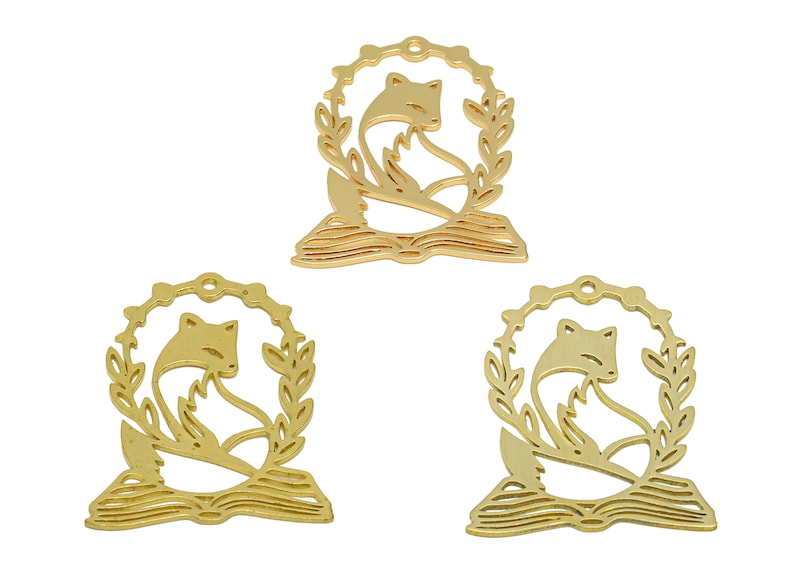 Pode incluir: Tr&ecirc;s pingentes de metal dourado com uma raposa enroscada em um livro com uma coroa de folhas ao redor.