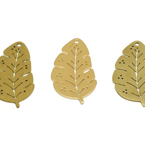 Puede incluir: Tres abalorios de metal dorado en forma de hoja con un diseño recortado. Las hojas tienen un patrón de nervaduras detallado y pequeños puntos.