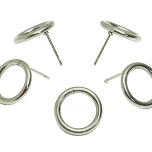 Puede incluir: Cinco pendientes de botón circulares de color plateado. Cada pendiente presenta un diseño de aro liso y redondeado sujeto a un poste para perforar. Los pendientes se muestran sobre un fondo blanco, resaltando su brillo metálico.