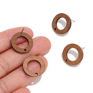 May include: Three circular brown wooden stud earrings, each featuring a central open hole and a small secondary hole near the edge. The earrings have a smooth, natural wood grain finish and include metal posts attached to the back for secure wear.