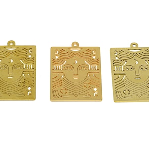 May include: Three gold-colored metal charms with a cutout design of a woman's face with a crescent moon above her forehead. The charms are rectangular and have a loop at the top for hanging.