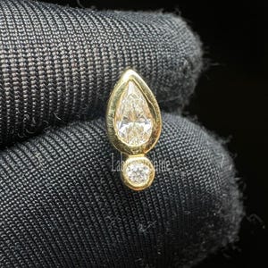 May include: A gold-colored earring with a pear-shaped diamond at the top and a smaller round diamond below. The diamonds sparkle against the dark background. The earring is a piece of jewelry.