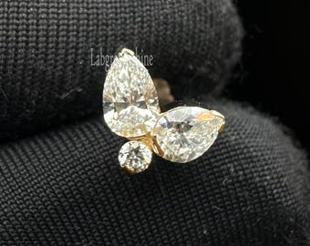 Puede incluir: Un pendiente dorado en forma de mariposa con tres piedras preciosas claras y brillantes. El pendiente presenta una piedra en forma de pera para cada ala y una piedra redonda para el cuerpo. La joya está sobre un fondo oscuro.