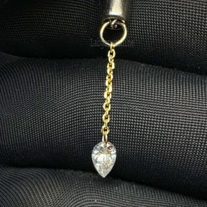 Puede incluir: Un pendiente de color dorado con un colgante de gema transparente en forma de pera. El pendiente está sujeto a una cadena y a un pequeño aro. El fondo es una tela oscura y texturizada.