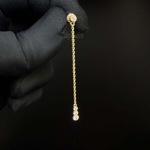 May include: A gold chain earring featuring a single round bezel-set diamond at the top post and a vertical drop chain ending in three small round bezel-set diamonds. The piece is crafted from yellow gold and displays a delicate, linear design.