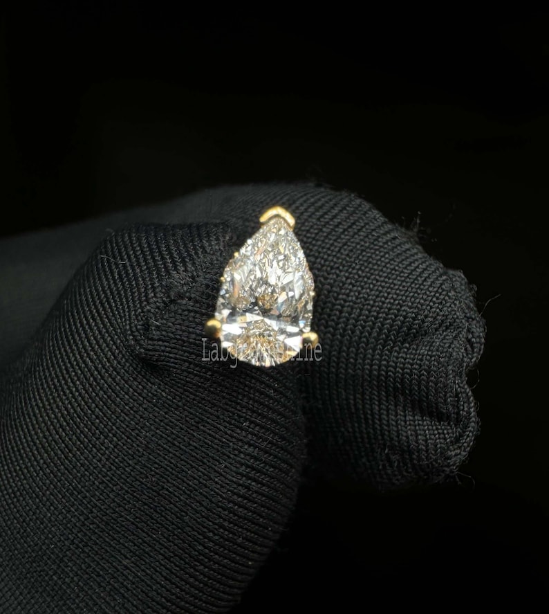May include: A pear-shaped diamond gemstone set in a gold setting. The diamond is clear and sparkly.