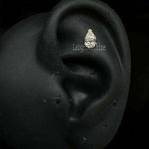 May include: A close-up of a clear pear-shaped gemstone set in a gold earring. The earring is worn in a model's ear.