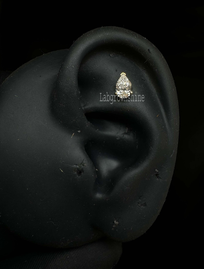 May include: A close-up of a clear pear-shaped gemstone set in a gold earring. The earring is worn in a model's ear.