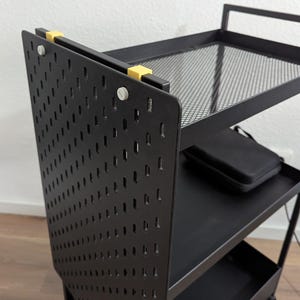 Op de afbeelding: Een zwarte metalen utility cart met drie planken. Het zijpaneel heeft een geperforeerd ontwerp. De bovenste plank is een gaas. Gele clips bevestigen het zijpaneel. Een zwarte koffer rust op de middelste plank.