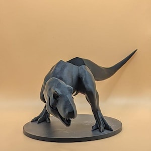 Op de afbeelding: Een zwart 3D-geprint Tyrannosaurus Rex dinosaurusbeeldje met een lange staart, staande op een ronde basis.