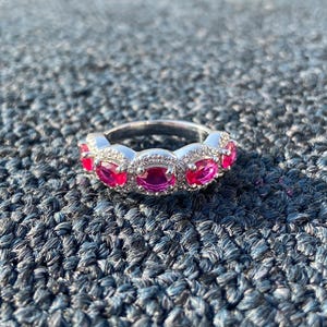 Peut inclure: Une bague en argent ornée de cinq pierres précieuses roses vives de forme ovale. Chaque pierre rose est entourée de petites pierres d'accent claires. La bague est posée sur une surface texturée bleu foncé.