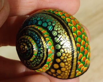 Puede incluir: Una concha de mar pintada a mano con un patrón colorido y en espiral de verde, azul, dorado y naranja. La concha está decorada con pequeños puntos y líneas.