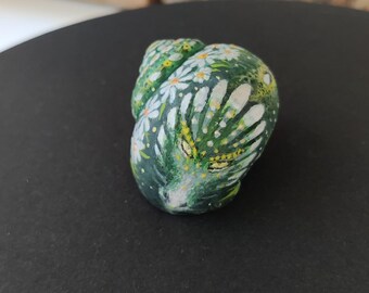 Puede incluir: Una piedra decorativa pintada a mano en tonos verdes, blancos y amarillos. La piedra está adornada con un patrón floral, con flores blancas en forma de margarita y hojas verdes. Se muestra sobre una superficie negra.