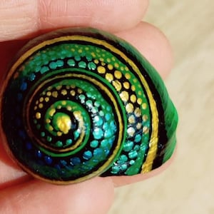 Puede incluir: Una concha de caracol pintada a mano con un patrón de espiral verde y dorado. La concha está pintada con un diseño detallado de puntos y remolinos.