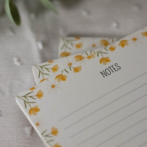 Puede incluir: Un bloc de notas blanco con un diseño floral con flores amarillas y hojas verdes. El bloc de notas tiene páginas rayadas y la palabra "NOTES" impresa en la parte superior.
