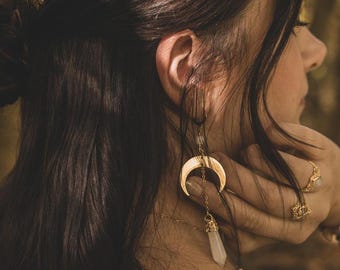 May include: Gold crescent moon earrings with a dangling crystal charm. The earrings are worn by a person with long brown hair.