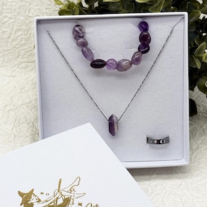 Könnte beinhalten: Ein Schmuckset in einer weißen Schachtel, darunter eine Halskette mit einem lila Kristallanhänger, ein Armband mit lila Perlen und ein silberner Ring mit einem schwarz-weißen Design. Eine weiße Karte mit goldenem Text befindet sich im Vordergrund.