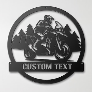 May include: A circular black metal wall sign featuring a silhouette of a person riding a motorcycle through a forest. The design includes a horizontal bar at the bottom with the words CUSTOM TEXT cut out in a bold, sans-serif font style.