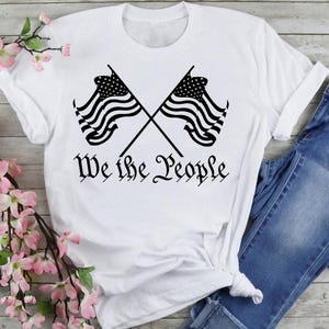 May include: White t-shirt with a black graphic of two crossed American flags and the text "We the People".
