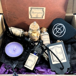 May include: A black box filled with a variety of witchy items, including a purple candle, a small bottle of essential oil, a black pouch, a small slate with a white circle design, and several small glass bottles with cork tops. The bottles are labelled "Apple", "Rainbow Moonstone", and "Lucky Charms Morgana".