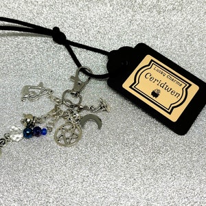 May include: A silver charm bracelet featuring a pentagram, crescent moon, and beads. It's attached to a black tag with "Lucky Charms Ceridwen". The background is a shimmering silver surface. The charms are attached to a silver-toned clasp.