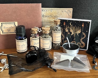 May include: A black obsidian crystal, a small black candle, three glass bottles with cork stoppers, a black pouch with a silver key charm, a silver charm bracelet with a key, a crescent moon, and a pentagram charm, a small bag of dried herbs, and a card with a black and gold illustration of three women with snakes. The bottles are labeled "Rose", "Mugwort", and "Lavender".