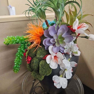 May include: A black vase with a variety of artificial plants and flowers. The vase is decorated with green moss and red flowers. The plants include succulents, air plants, and a spiky orange flower.