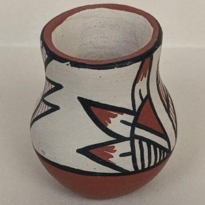May include: A small, hand-painted pottery vase with a white background and geometric designs in black, red, and white. The vase has a rounded body and a wide opening.