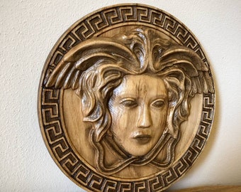 Op de afbeelding: Een houten wanddecoratie met een gesneden afbeelding van een vrouwenhoofd met een gevleugeld hoofdtooi, omringd door een Grieks sleutelmotief.