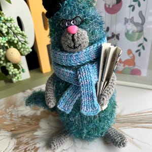 May include: A handmade, teal cat figurine wearing a black top hat, round glasses, and a blue and purple scarf. The cat holds a newspaper and has a pink nose. A whimsical, handcrafted cat ornament.
