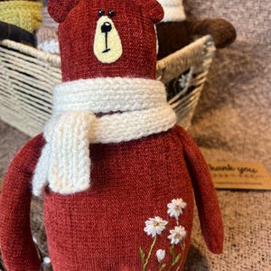May include: A red knitted teddy bear with a white knitted scarf and embroidered white flowers. The bear is sitting in a wicker basket.