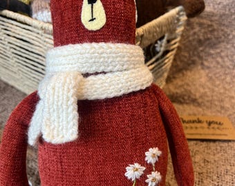 May include: A red knitted teddy bear with a white knitted scarf and embroidered white flowers. The bear is sitting in a wicker basket.