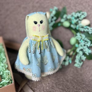 以下が含まれることがあります： 手作りの黄色のウサギのぬいぐるみで、青いドレスを着ています。ドレスには黄色のリボンと白いレースのトリムが付いています。ぬいぐるみは茶色の表面に置かれており、背景には緑と白の葉があります。