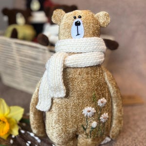 May include: A plush brown bear wearing a white knitted scarf and embroidered with white flowers. The bear is sitting on a light brown surface.